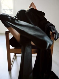 Model in black coat and gloves seated in a wooden armchair, leaning back with legs raised toward camera in a minimalist interior room.
