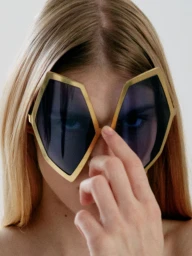 Close-up of a blonde woman wearing geometric gold-rimmed sunglasses and holding her hand up to the frame.