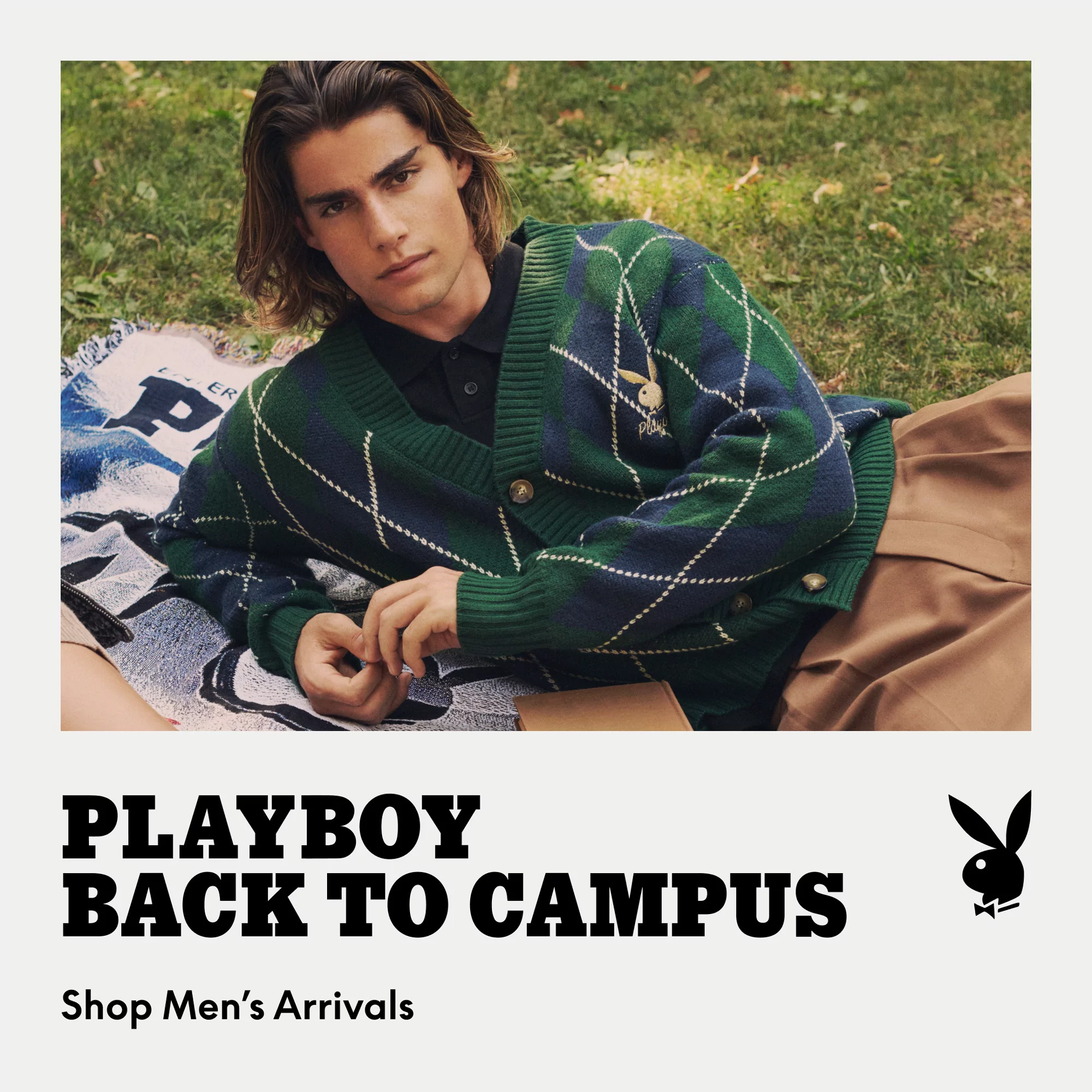 A young adult male model wearing a green plaid cardigan sweater lies on a blanket in a grassy outdoor setting for a fashion advertisement.