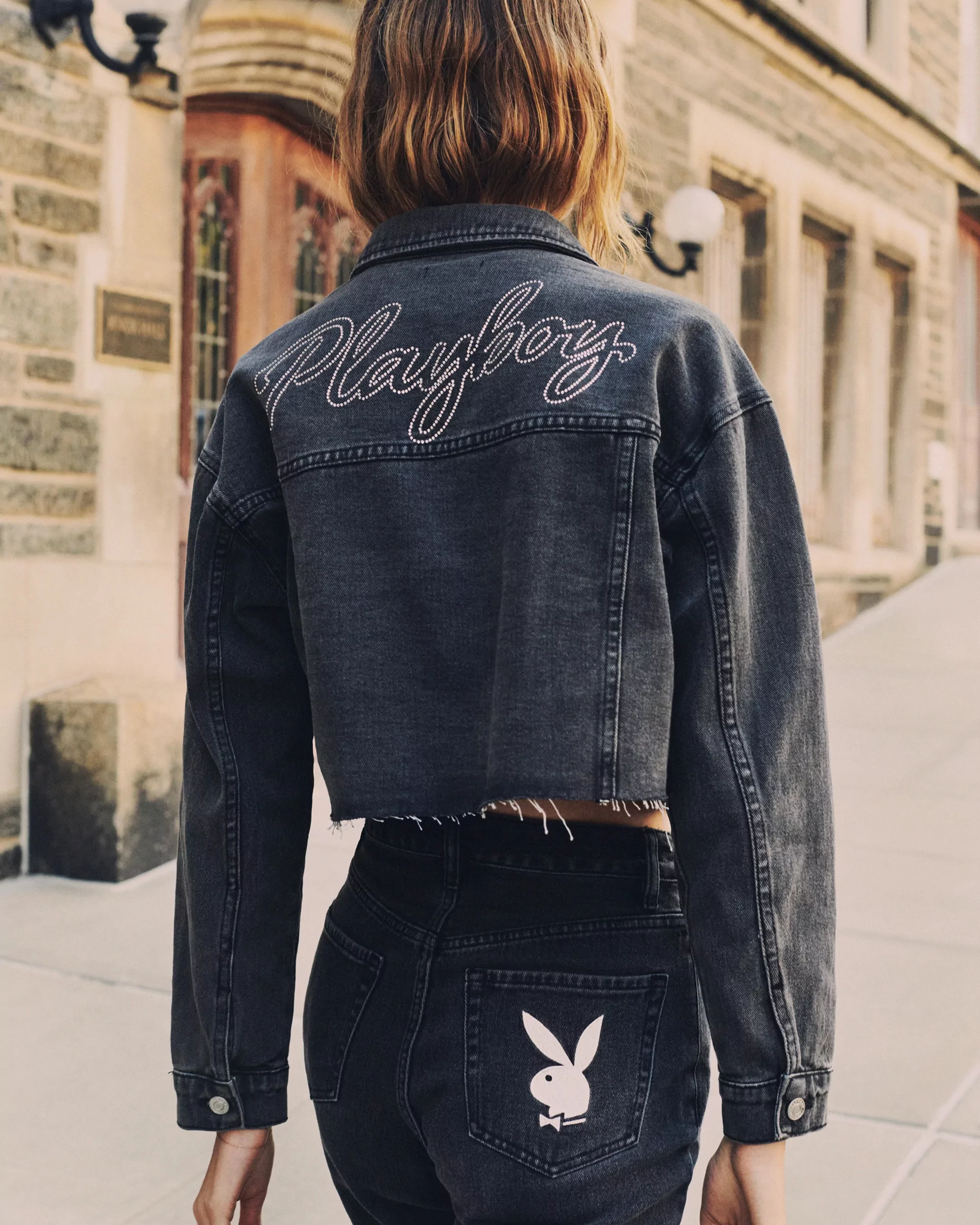 Back view of a woman in a gray denim jacket and dark jeans featuring a Playboy bunny logo, standing outdoors on a paved area.