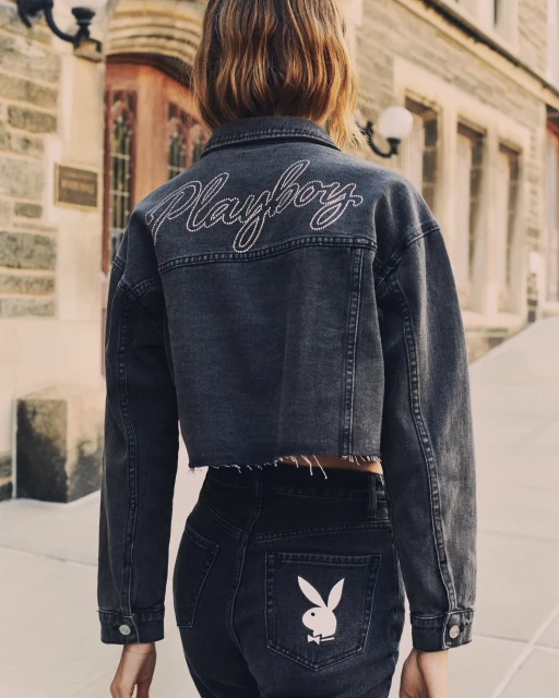 Back view of a woman in a gray denim jacket and dark jeans featuring a Playboy bunny logo, standing outdoors on a paved area.