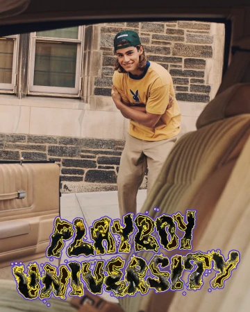A young adult man with long hair and a baseball cap stands in front of a stone building, wearing a logo t-shirt inside a car.