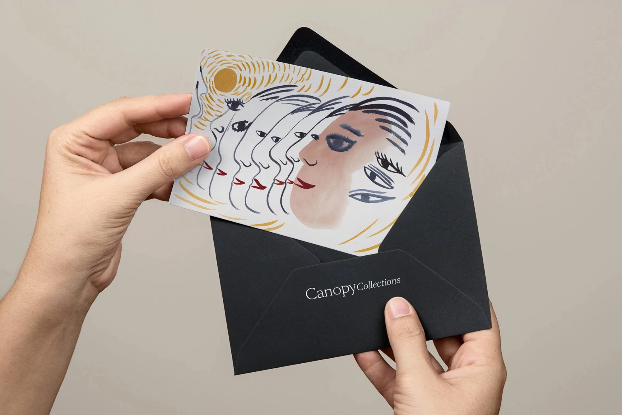 Hands holding an envelope containing a card with an illustration of multiple stylized faces, on a neutral background in soft lighting.