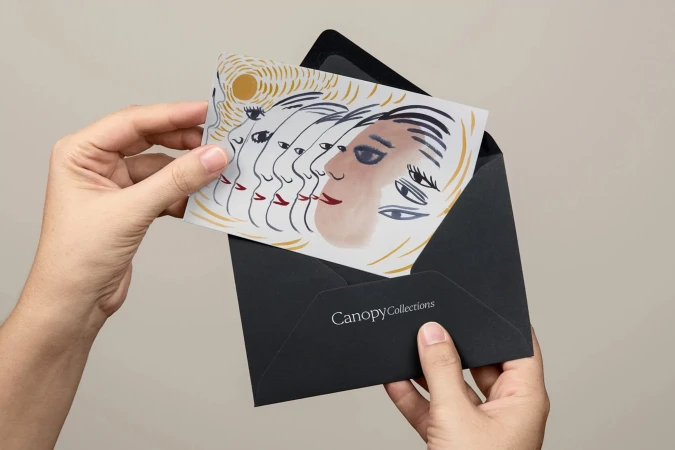 Hands holding an envelope containing a card with an illustration of multiple stylized faces, on a neutral background in soft lighting.