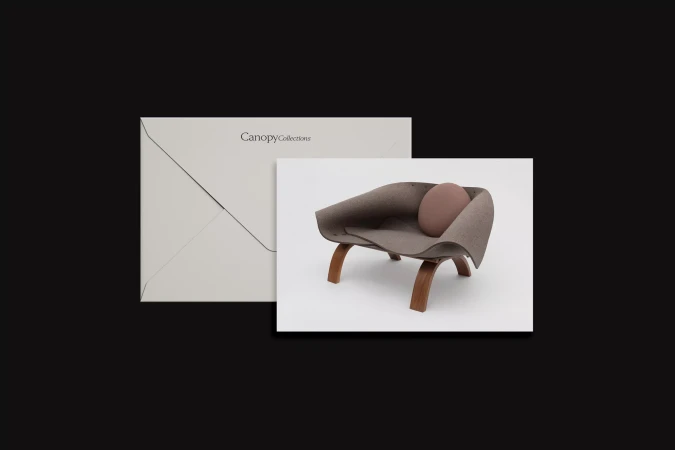 Studio shot of a brown upholstered modern chair with wood frame and a round pillow next to a light grey envelope on dark grey.