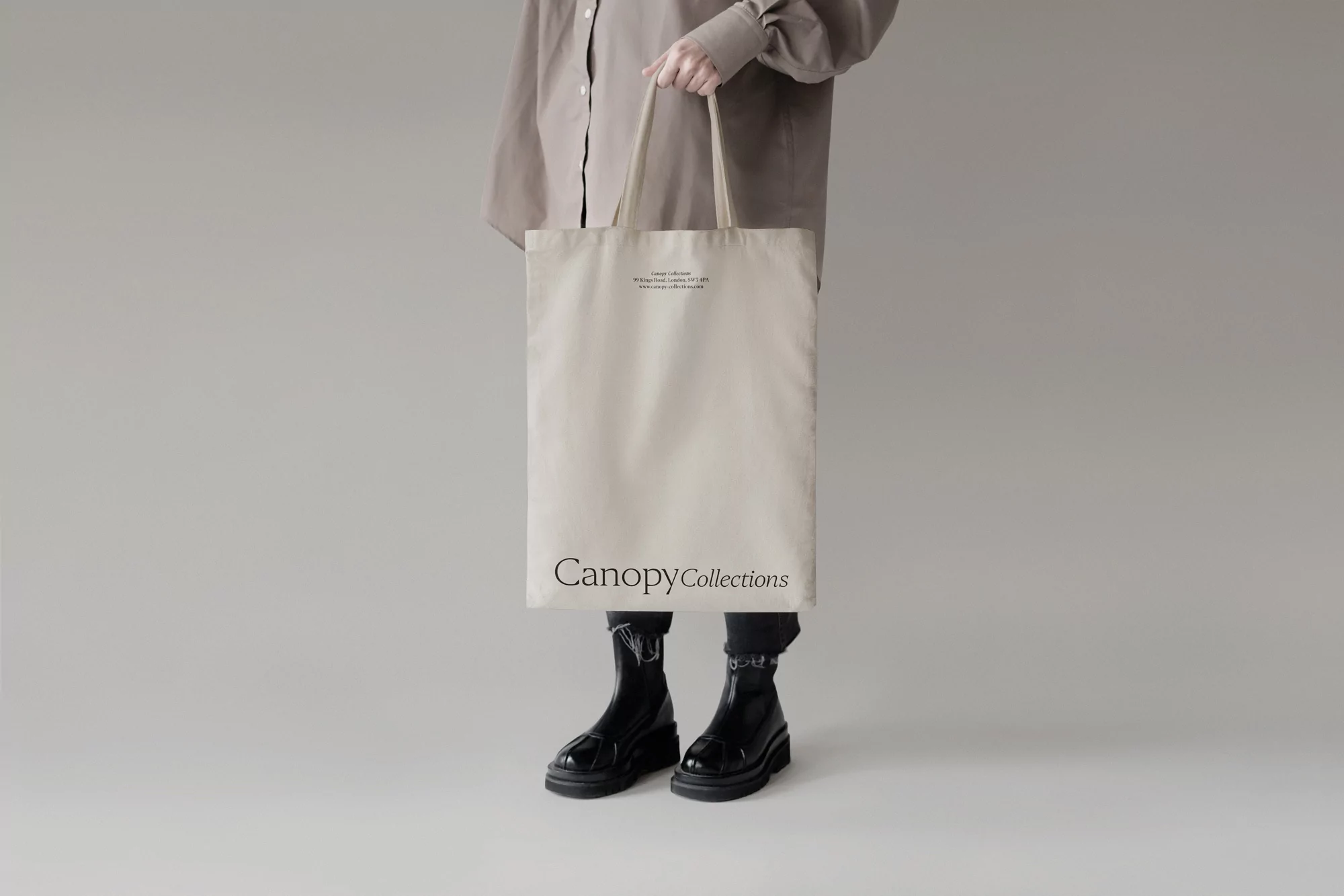 Person holding a canvas tote bag in a studio. The bag has black text on the front and is paired with a neutral-colored shirt and black boots.