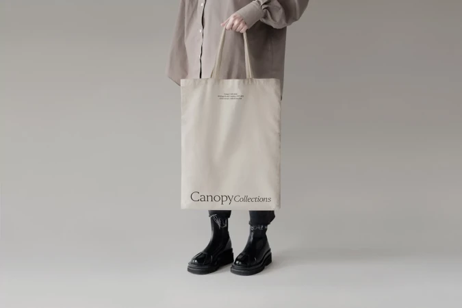 Person holding a canvas tote bag in a studio. The bag has black text on the front and is paired with a neutral-colored shirt and black boots.