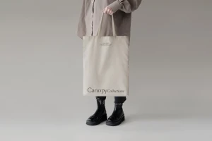 Person holding a branded canvas tote bag wearing a beige shirt and black boots against a plain grey backdrop.