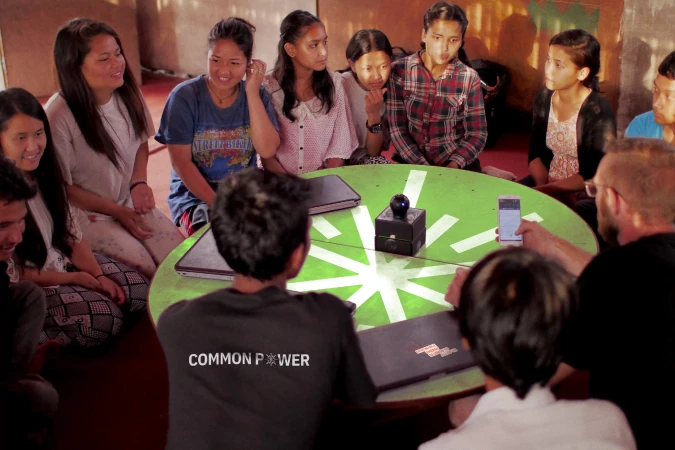 Group of teenagers gather around a table with electronic devices, engaging in discussion, captured in an indoor setting.