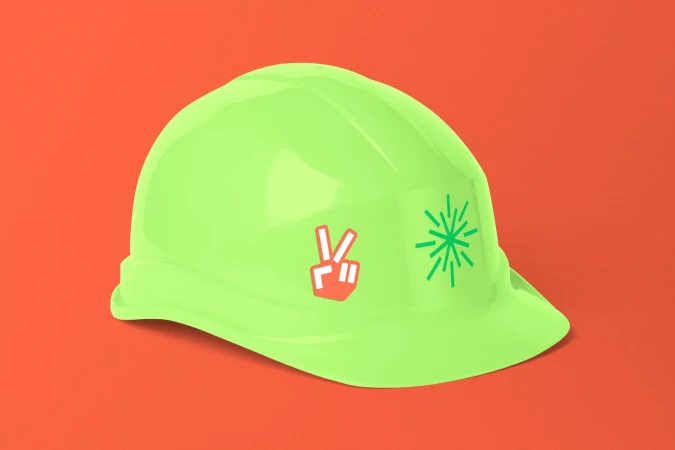 Bright neon green construction hat with peace sign and starburst stickers on a vivid orange background with subtle shadows.