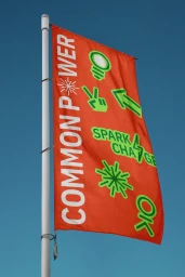 Bright orange flag with white Common Power branding and green graphic icons flying against a clear blue sky.