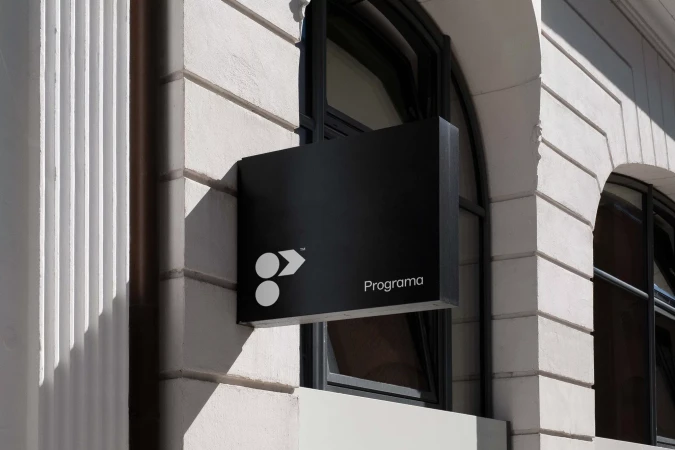 Exterior daytime shot of a black business sign for Programa mounted on a white building facade with arched windows visible behind it.
