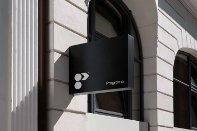 Black rectangular sign with white Programa logo mounted on a textured white building exterior wall next to a dark-framed arched window.