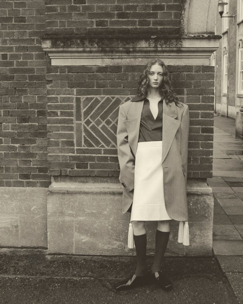 Monochrome portrait of a woman in a skirt and coat, standing against a brick wall in an outdoor setting with natural lighting.