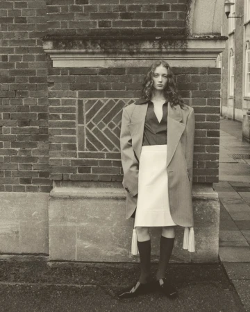 Monochrome portrait of a woman in a skirt and coat, standing against a brick wall in an outdoor setting with natural lighting.