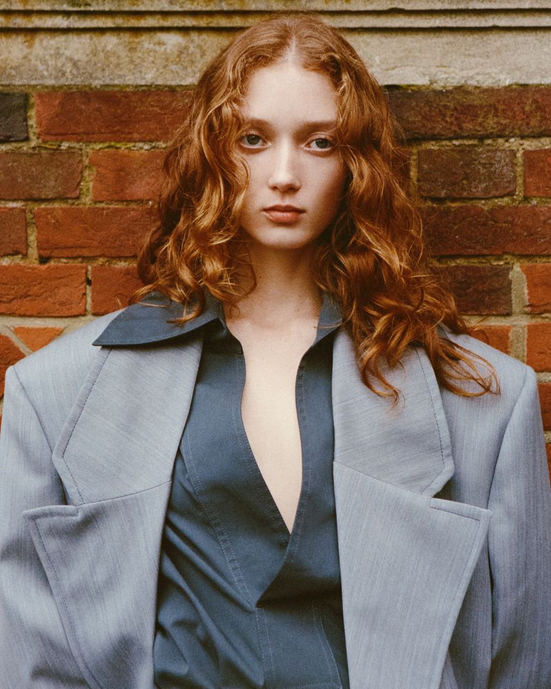 Portrait of a red-haired woman with fair skin wearing a gray blazer and blue top, posing in front of a brick wall in natural lighting.