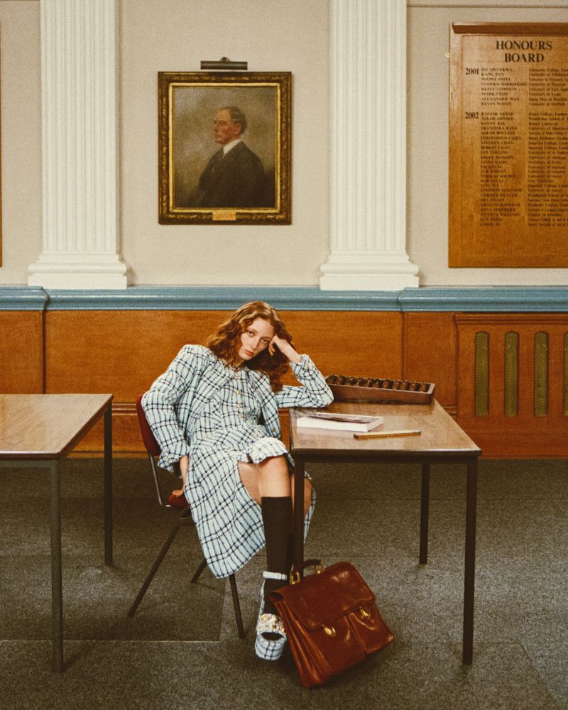 Young woman in plaid dress sits at school desk with bag, styled in platform shoes and knee-high socks in front of portrait.