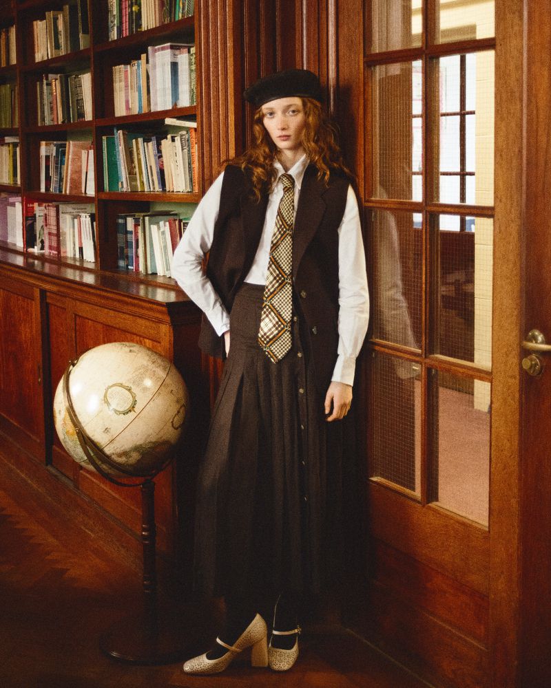 Young woman in preppy fashion poses indoors by bookshelves wearing vest, tie, skirt, and Mary Jane heels.