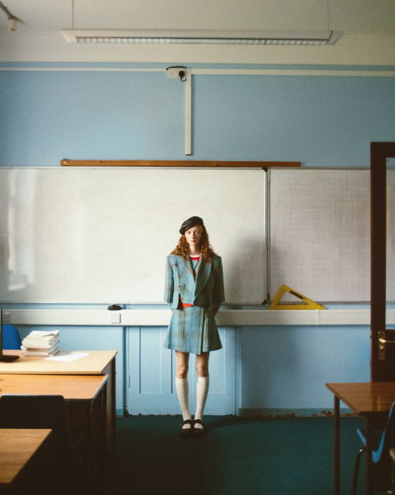 Young woman in a school uniform stands in a classroom with desks and a whiteboard, creating an editorial fashion style image.