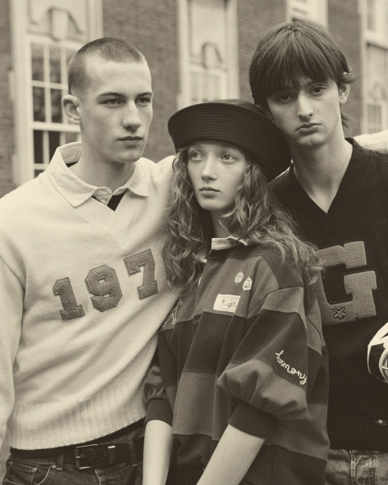 Three models, two males and one female, pose in front of a brick building, wearing vintage-style tops and displaying neutral expressions.