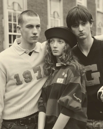 Three models, two males and one female, pose in front of a brick building, wearing vintage-style tops and displaying neutral expressions.