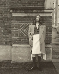 Model standing against an exterior brick wall wearing an oversized coat, white skirt, and dark knee-high socks.