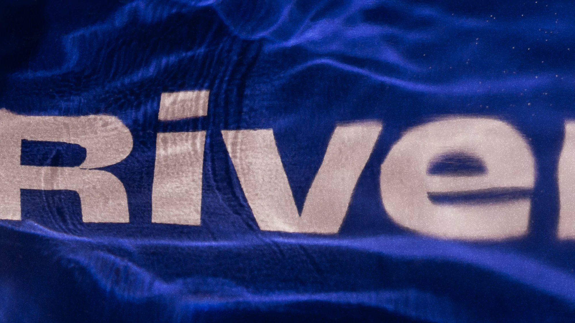 Close-up abstract of the word 'River' in white lettering against a textured, dark blue background evoking rippling water.