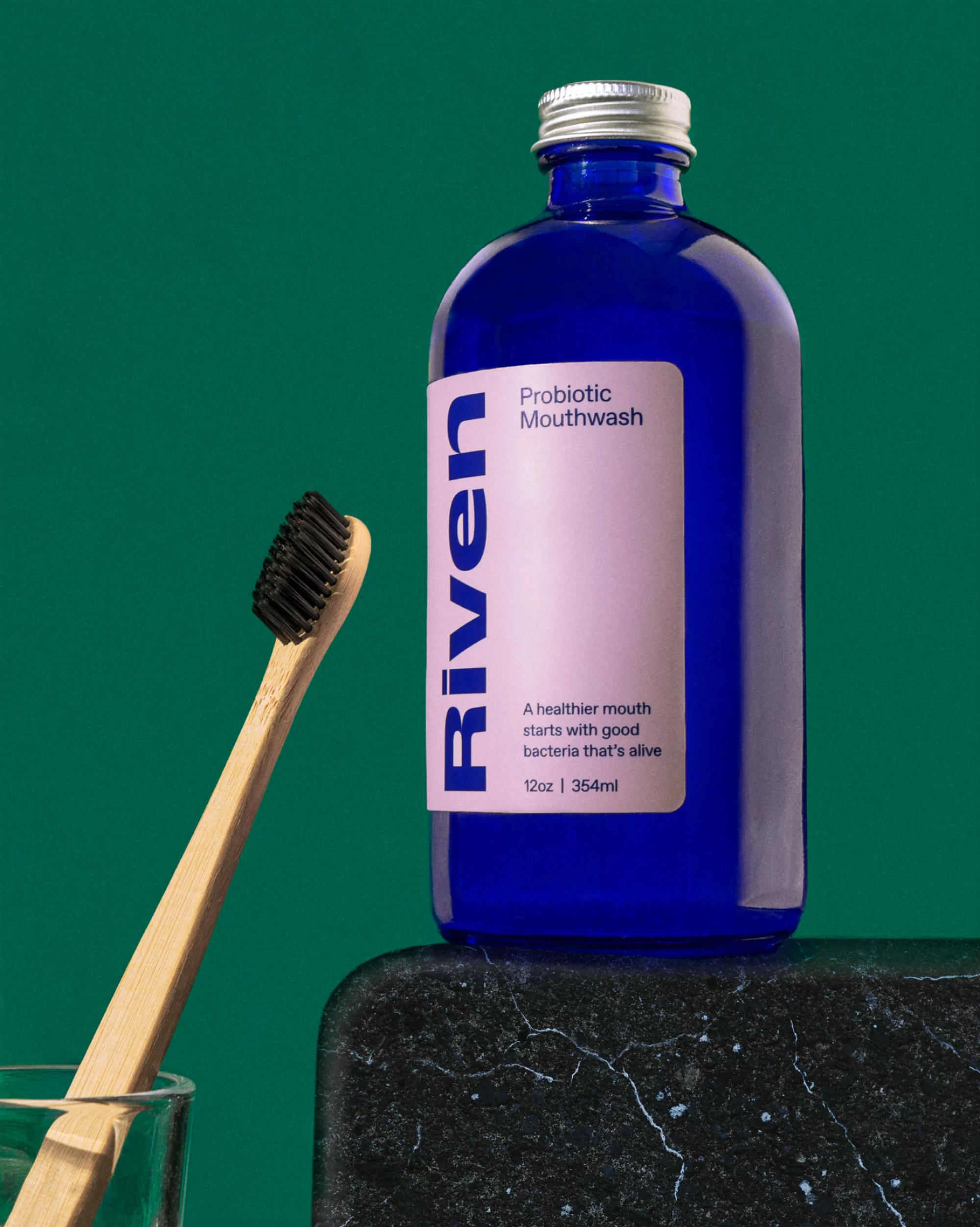 Probiotic mouthwash in a blue bottle with a silver cap, placed on a black marble surface with a toothbrush in a glass.