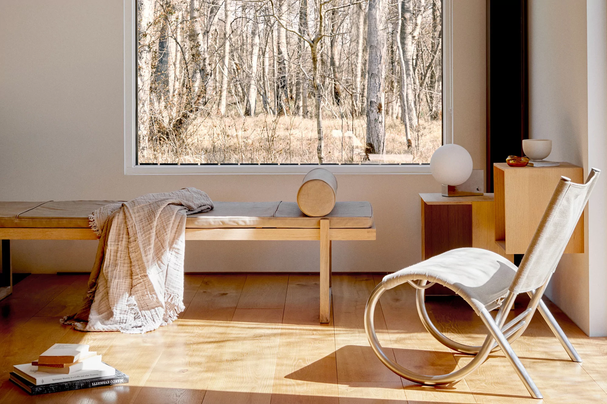 An interior room scene displays a light wooden daybed with throw, a curved chair, a white spherical lamp, and a view of a forest through a window.