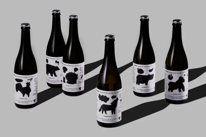 Seven black bottles of Quadraft Brewing beer with animal label art, set on a gray background with natural shadows.