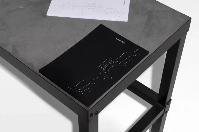 Black book with dotted line art on a dark grey table, next to a white booklet with similar dotted design, against a white background.