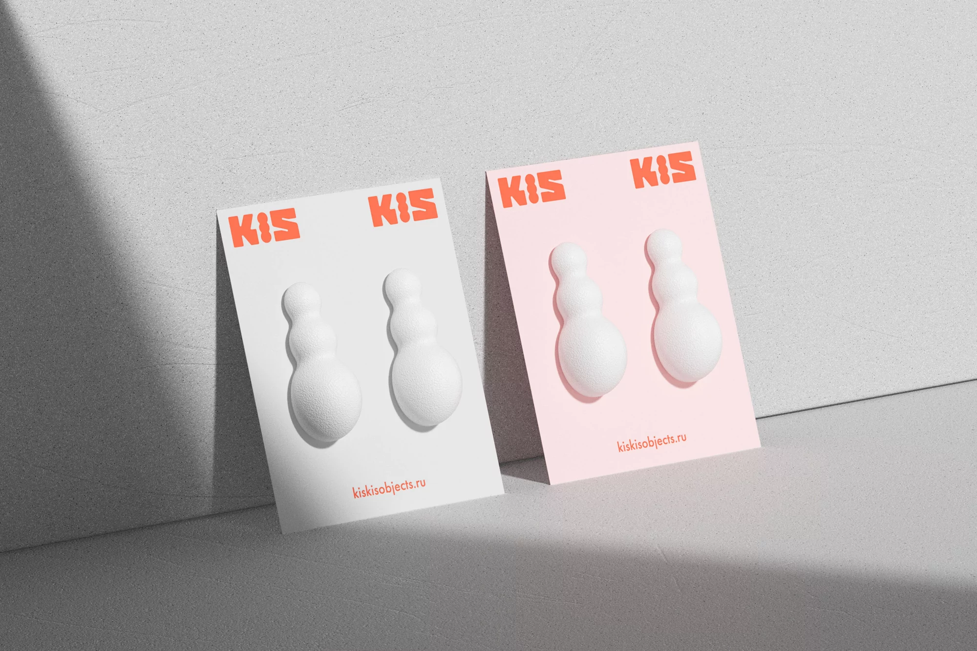 Close-up of pair of white, spherical earrings on a pink and white display card from kisikisobjects.ru, against a grey wall backdrop.