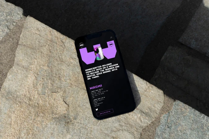 Smartphone displaying Wagmi beer website on a stone surface with natural light. Black mobile phone and purple beer brand.