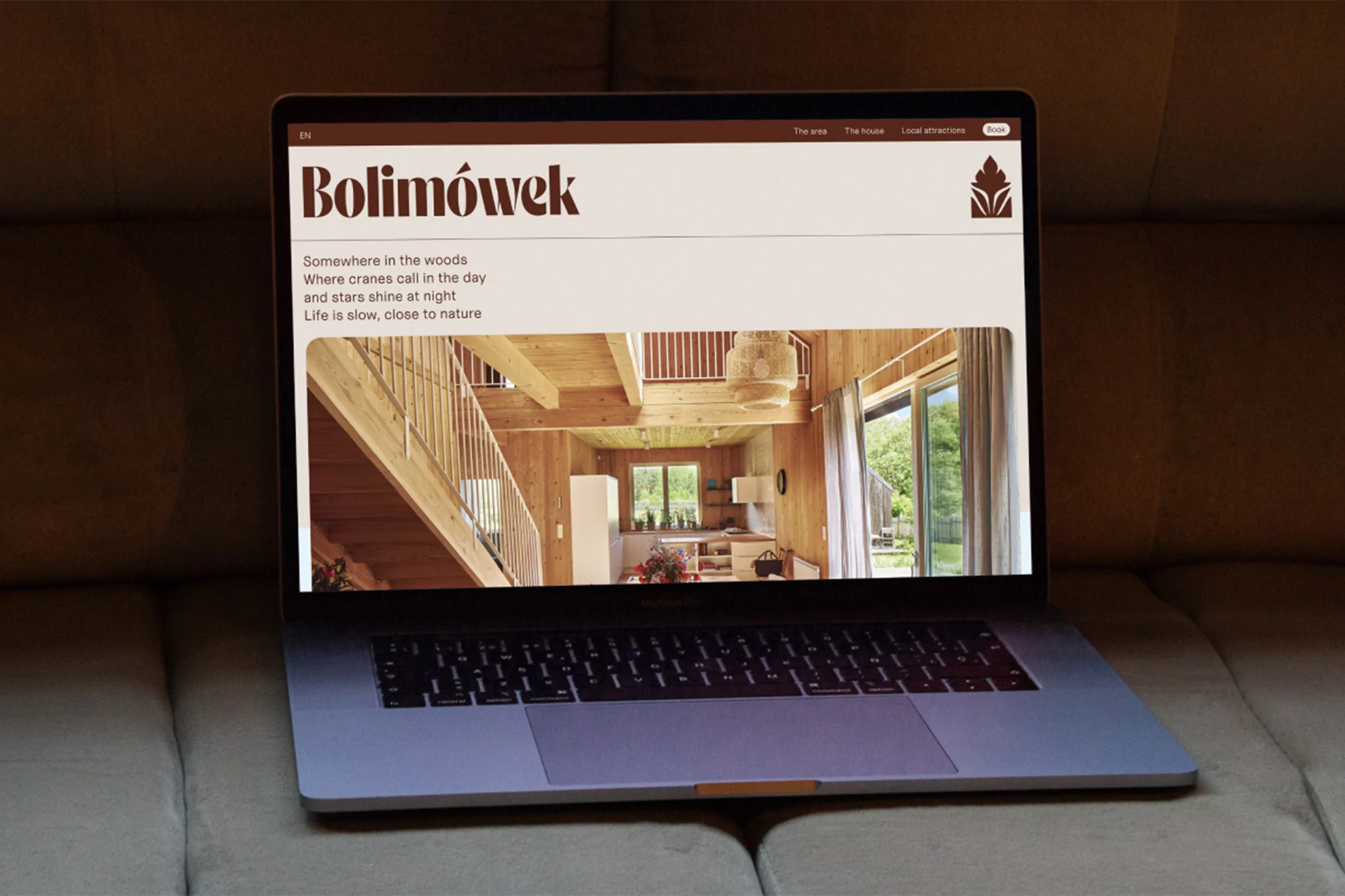 Laptop displaying a Bolimowek website showcasing a wooden house interior, set on a beige sofa, creating an inviting, tech-meets-rustic scene.