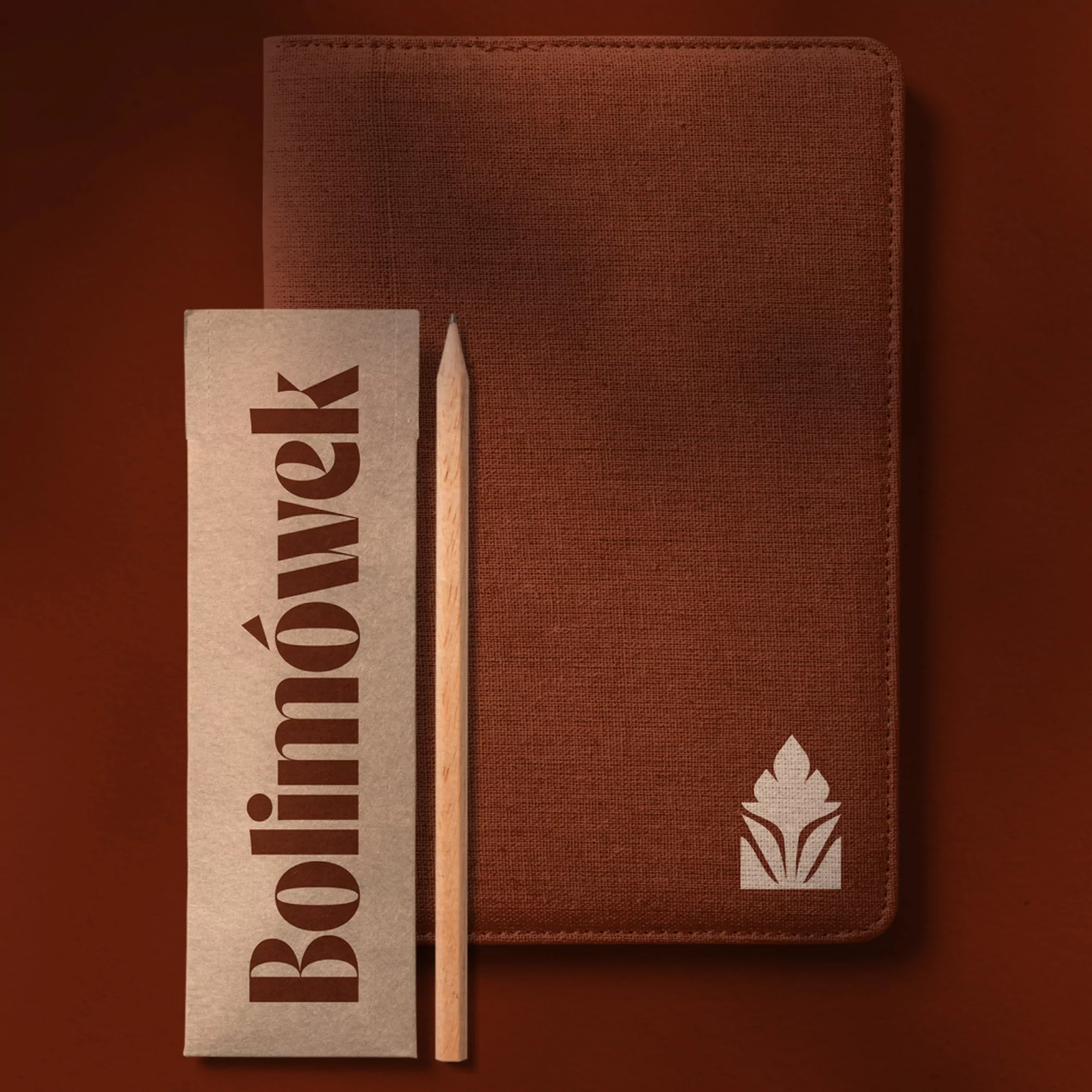 Brown notebook with a logo, a wooden pencil, and a branded paper card on a plain brown background. Stationery flat lay.