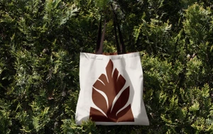 White tote bag with brown leaf graphic hanging against a dense background of green pine tree foliage
