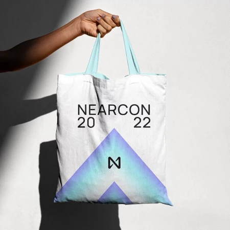 Hand holds a white tote bag with light blue straps displaying the text 'NEARCON 2022' and a geometric logo against a white wall.