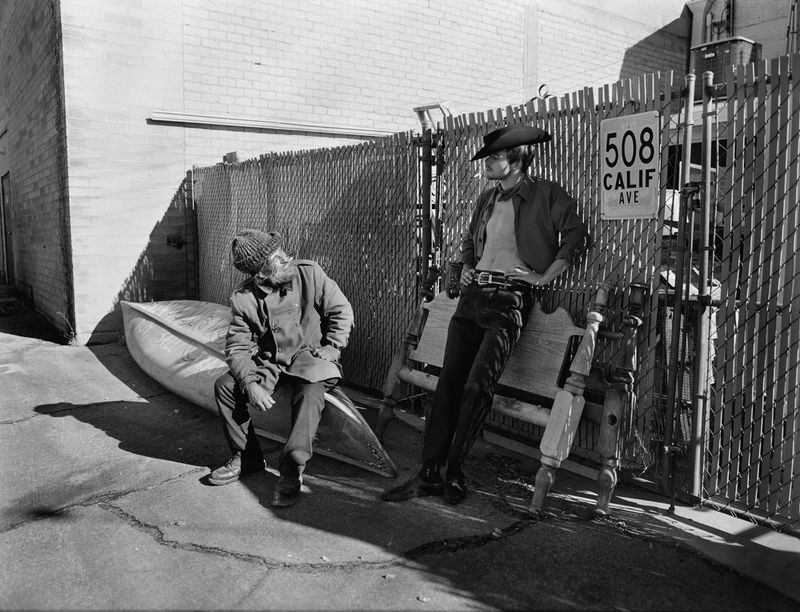 Two men pose in an outdoor black and white portrait featuring a cowboy hat, a canoe, and a metal fence along a city street.