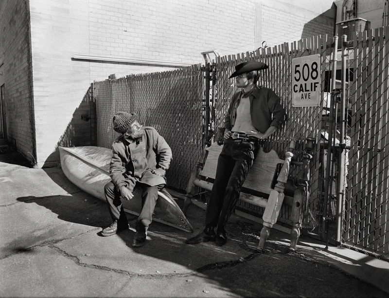 Two men pose in an outdoor black and white portrait featuring a cowboy hat, a canoe, and a metal fence along a city street.