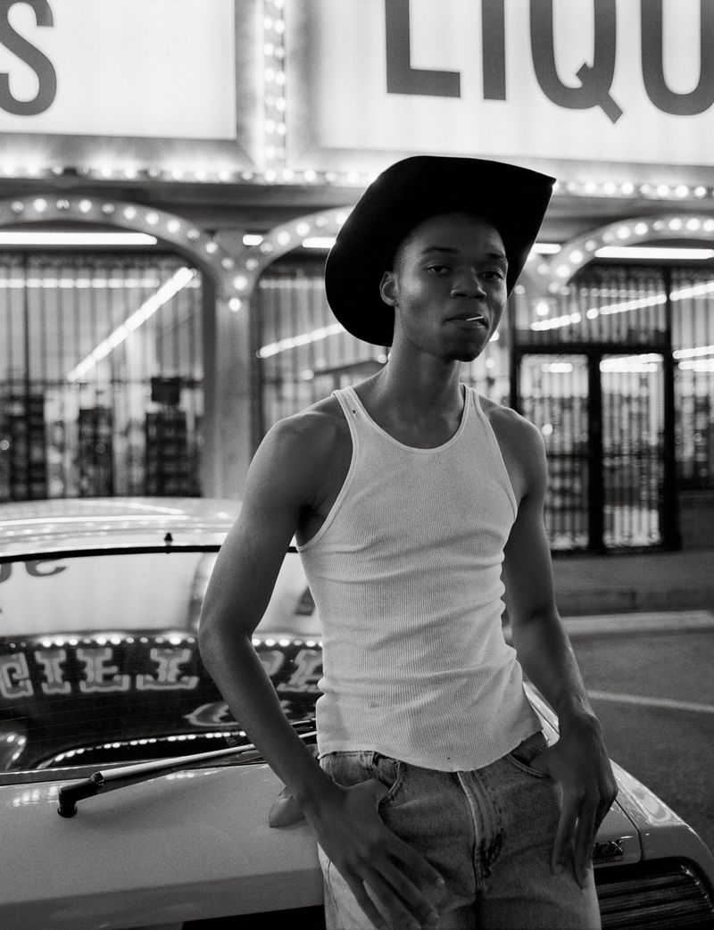 Black and white portrait of a Black man with a cowboy hat and cigarette leaning on a vintage car in front of a liquor store.