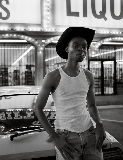 Black and white portrait of a Black man with a cowboy hat and cigarette leaning on a vintage car in front of a liquor store.