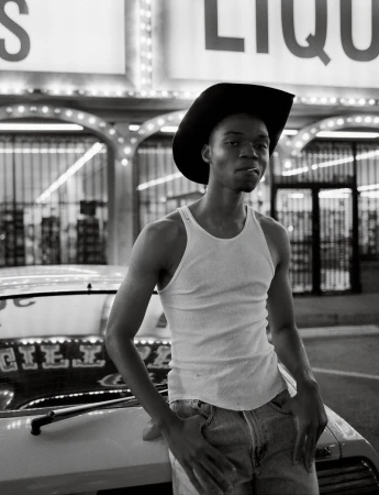 Black and white portrait of a Black man with a cowboy hat and cigarette leaning on a vintage car in front of a liquor store.
