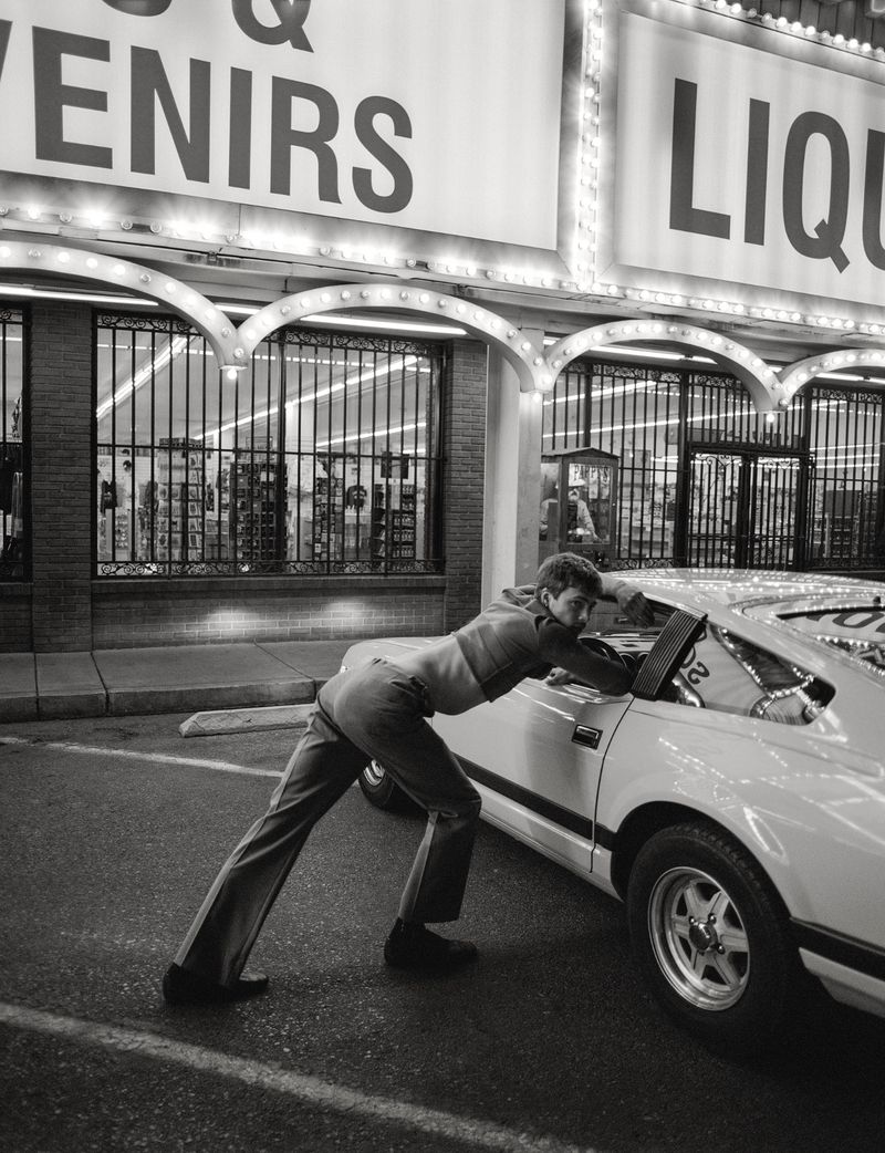 Monochrome photo of an adult male pushing a car in front of a store with lit signs in an outdoor parking lot setting at night.