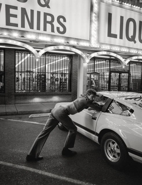 Monochrome photo of an adult male pushing a car in front of a store with lit signs in an outdoor parking lot setting at night.