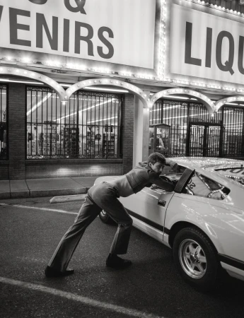 Monochrome photo of an adult male pushing a car in front of a store with lit signs in an outdoor parking lot setting at night.