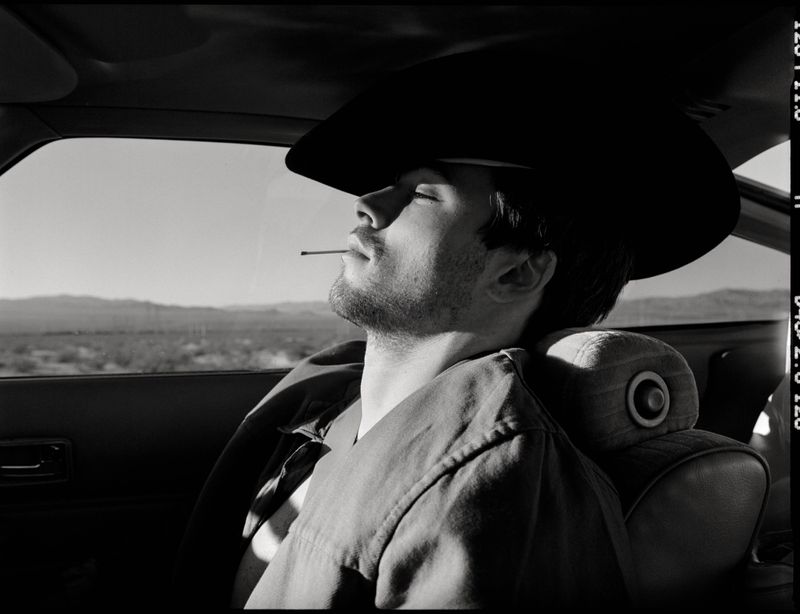 Monochrome portrait of a man in a cowboy hat, reclining in a car seat with a toothpick in his mouth, set against a desert backdrop.