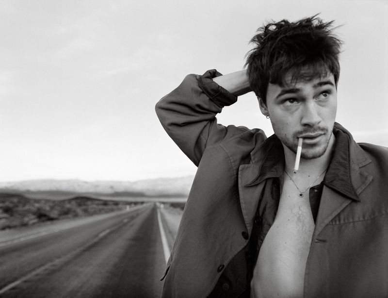 Black-and-white portrait of a young adult man in an open jacket with a cigarette in his mouth, standing on a long road.
