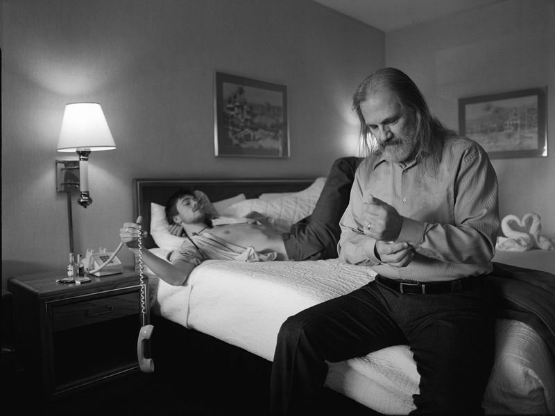 Black-and-white shot inside a motel room featuring two adult men, one sitting on the edge of the bed and the other laying on it.