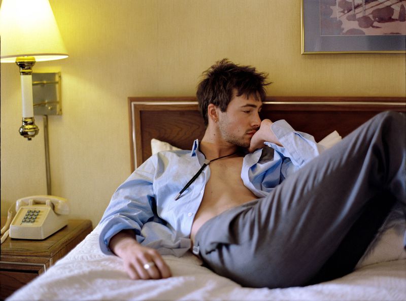 Adult male in an unbuttoned shirt and dress pants reclines on a bed, with a telephone and lamp visible in the hotel room setting.