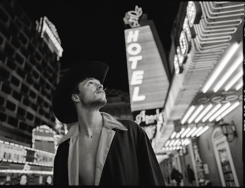 A young adult male in a cowboy hat stands on a city street at night, looking up at illuminated hotel signs and building facades, in black and white.
